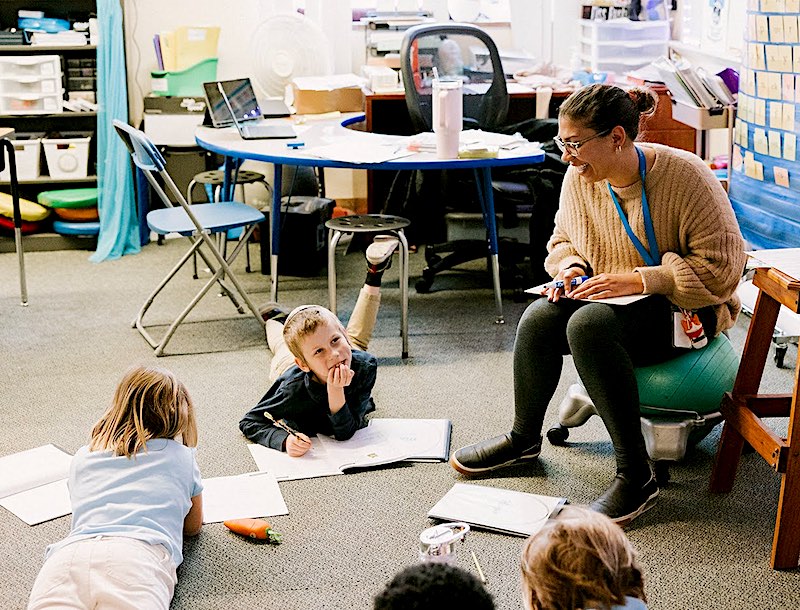 “students at PCS West 1st-5th grade tuition-free charter writing in notebooks, laying on the floor of their classroom talking with their teacher”