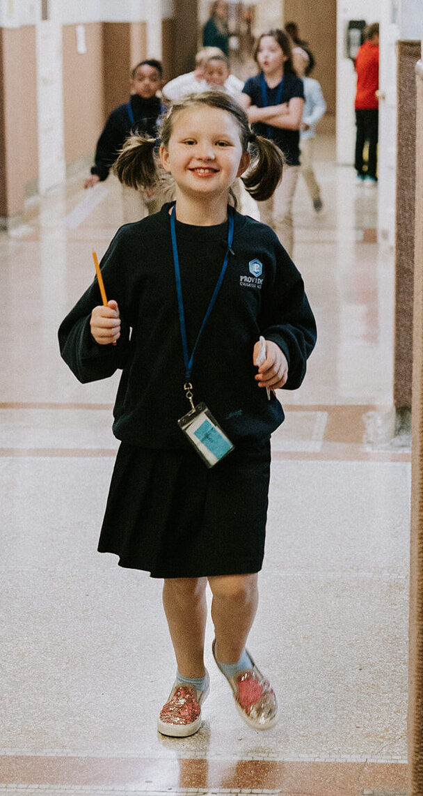 A smiling young girl walks in a school hallway with a pencil in one hand. Her hair is in pigtails and she is wearing a blue school uniform.