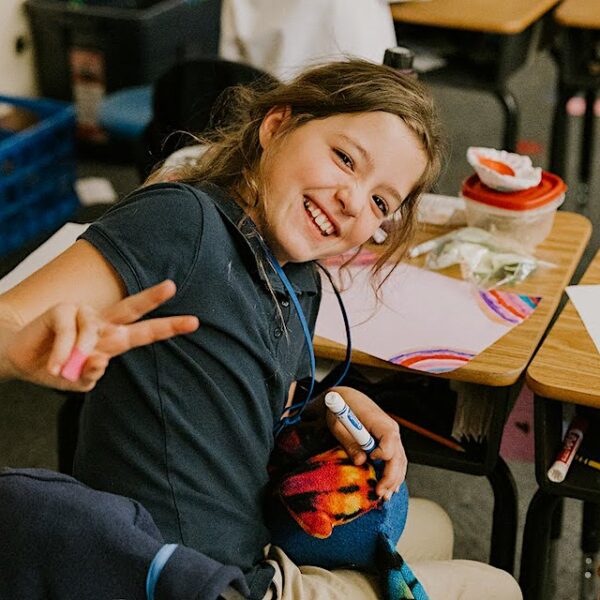 Student at PCS West tuition-free 1st-5th charter school smiling and holding up two fingers in a peace sign while sitting at her desk.